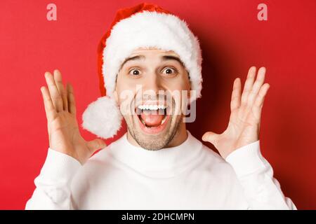 Close-up of happy young man in santa hat, making big christmas announcement, smiling amazed, standing over red background Stock Photo