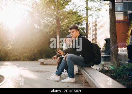 Full length of friends using mobile phone while sitting on retaining wall Stock Photo