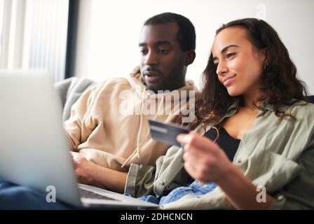 Young mixed ethnicity couple sit side by side on sofa at home holding credit card and shopping online using laptop Stock Photo