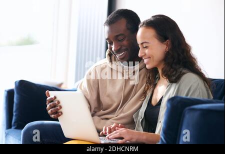 Young mixed ethnicity couple sit side by side on sofa at home browsing internet on laptop Stock Photo