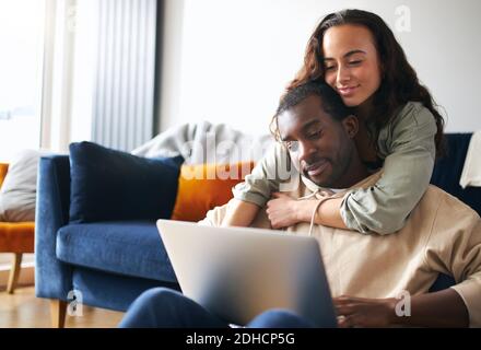 Woman hugging man from behind as young mixed ethnicity couple relax on sofa at home browsing internet on laptop Stock Photo