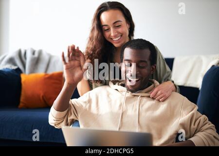 Woman sitting behind man as young mixed ethnicity couple relax on sofa at home making video call on laptop Stock Photo