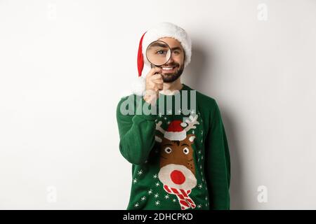 Christmas and holidays concept. Happy bearded man in Santa hat and xmas sweater, looking through magnifying glass and smiling, s Stock Photo
