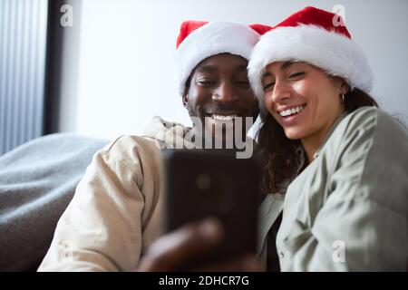 Young mixed ethnicity couple relaxing on sofa at home wearing santa hats posing for christmas selfie on mobile phone Stock Photo