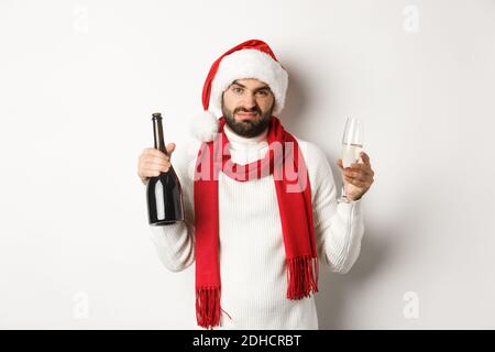 Christmas party and holidays concept. Skeptical bearded man in Santa hat and scarf, holding champagne and complaining, standing Stock Photo
