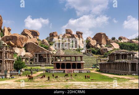 Hampi, Karnataka, India - November 4, 2013: Nandi Monolith Statue temple and nearby area. Cattle grazing in front. Ruinous buildings and big boulders Stock Photo