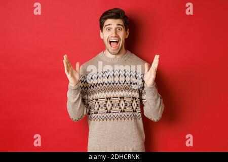 Portrait of surprised and happy handsome man, wearing christmas sweater, looking amazed, celebrating new year, standing over red Stock Photo