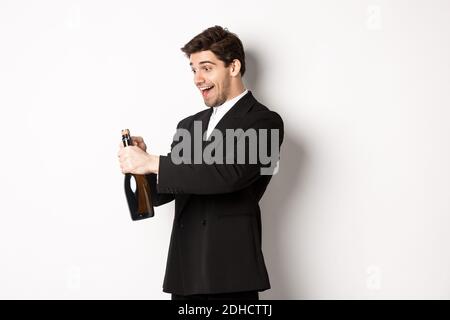 Portrait of attractive man in black suit, winking at camera and opening bottle of champagne, celebrating new year, standing agai Stock Photo