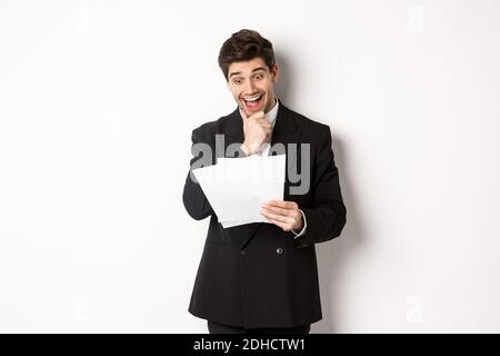 Portrait of handsome businessman looking excited at documents, working, standing against white background in black suit Stock Photo