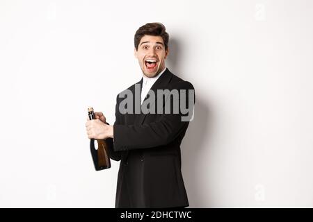 Portrait of attractive man in black suit, winking at camera and opening bottle of champagne, celebrating new year, standing agai Stock Photo