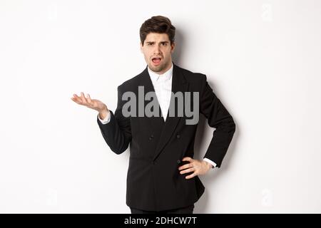 Portrait of arrogant man in black suit, looking confused and disappointed, complaining about something strange, standing over wh Stock Photo