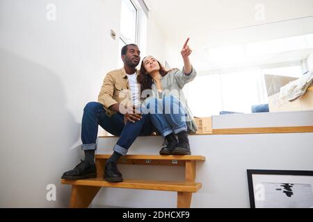 Young mixed ethnicity couple celebrating moving day in lounge of new home sitting on stairs surrounded by removal boxes together Stock Photo