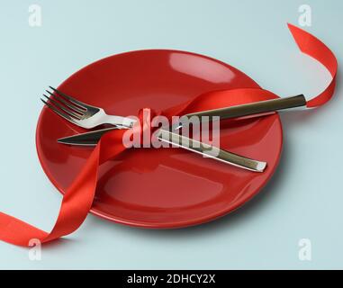 Round plate and metal fork and knife tied with a red silk ribbon, blue wooden background Stock Photo