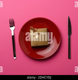 Gift box lies in a round ceramic plate and fork with a knife on a pink background Stock Photo