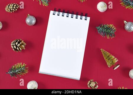 Empty notebook surrounded by seasonal decoration like golden fir cones, fir branches and Christmas tree baubles on dark burgundy background Stock Photo