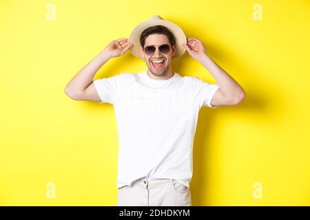 Happy man on vacation, wearing straw hat and sunglasses, smiling while standing against yellow background Stock Photo