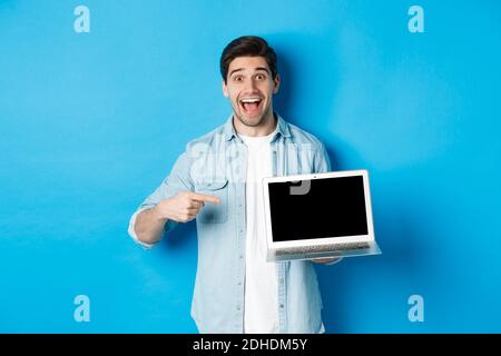 Excited smiling man pointing at laptop screen, showing internet promo offer or website advertisement, standing over blue backgro Stock Photo