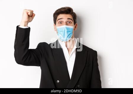 Concept of covid-19, business and social distancing. Close-up of excited businessman in medical mask and suit, rejoicing, achiev Stock Photo