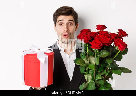 Close-up of attractive man in suit looking surprised, holding gift box and bouquet of roses, giving present for holiday, standin Stock Photo