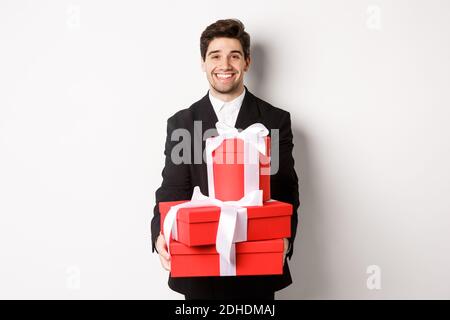Portrait of handsome bearded man in trendy suit, holding gifts for new year and smiling, prepared presents, standing over white Stock Photo