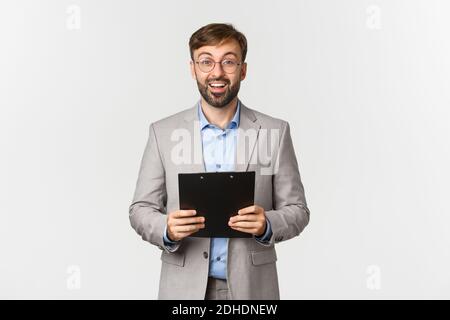 Image of confident surprised and happy businessman in grey suit and glasses, holding clipboard and looking at camera, standing o Stock Photo