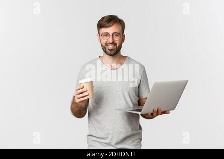 Portrait of handsome male freelancer, drinking coffee and working with laptop, standing over white background Stock Photo