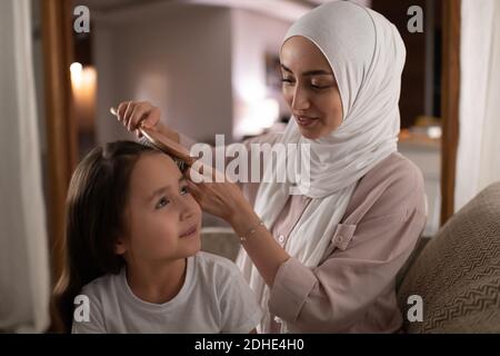 Adult Muslim woman smiling and combing hair of happy girl while sitting on sofa Stock Photo