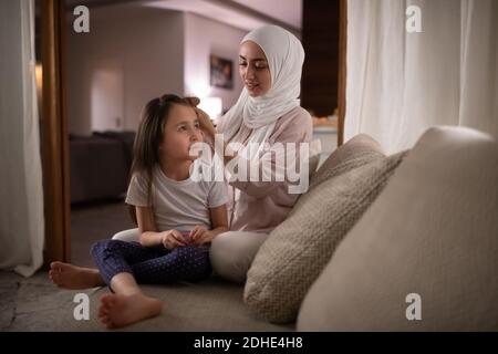 Happy Muslim mother combing hair of dreamy daughter on couch in cozy room at home Stock Photo