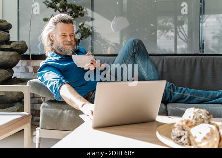 Mature man with a beard smiling and working with his laptop on a sofa Stock Photo