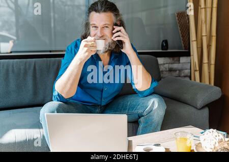 Mature man with beard talking on the phone and drinking coffee sitting Stock Photo
