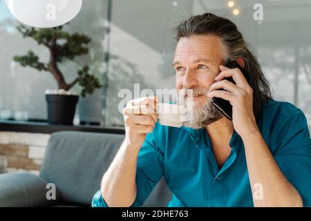 Mature man with beard talking on the phone and drinking coffee sitting Stock Photo