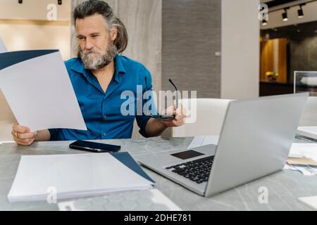 Bearded mature man reviewing some documents at his desk in front of his laptop at home. business concept Stock Photo