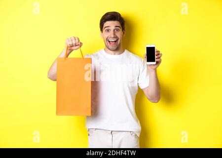 Surprised man holding shopping bag and showing smartphone screen, concept of mobile banking and app achievements, yellow backgro Stock Photo