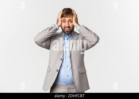 Portrait of tensed and distressed businessman with beard, wearing grey suit, holding hands on head and looking anxious or perple Stock Photo