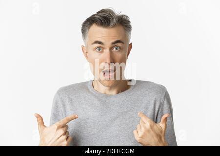 Close-up of confused middle-aged man, pointing at himself and gasping, standing over white background Stock Photo