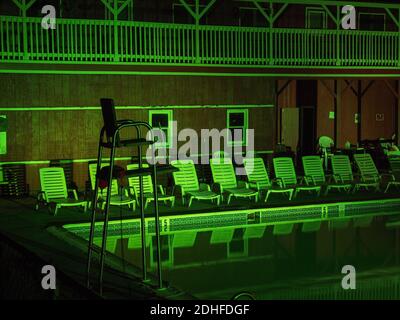 An empty swimming pool after dark is bathed in eerie green light outside a cedar cabin style pool house, surrounded by neatly lined up deck loungers r Stock Photo