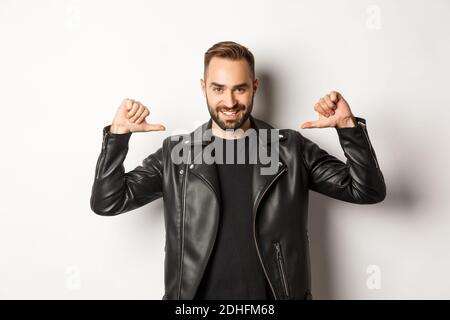 Confident handsome man wearing black leather jacket, pointing at himself and smiling self-assured, standing against white backgr Stock Photo