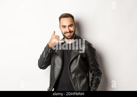 Handsome man in leather jacket flirting with you, showing mobile call me gesture, standing against white background Stock Photo