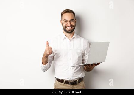 Business. Young successful male entrepreneur showing thumb-up while working on laptop, standing over white background Stock Photo
