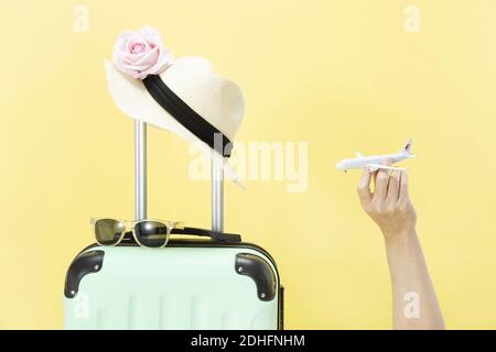 A closeup of a hand holding a toy airplane, sunglasses, hat, and luggage on a yellow background-concept travel Stock Photo
