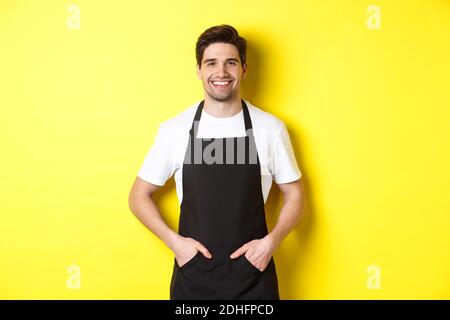 Happy barista in black apron looking at camera. Coffee shop owner wearing cafe uniform and smiling, standing over yellow backgro Stock Photo