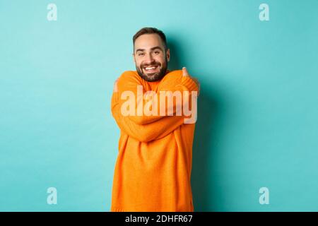 Happy man feeling comfortable, wearing warm sweater and hugging himself, smiling satisfied, standing over light blue background Stock Photo