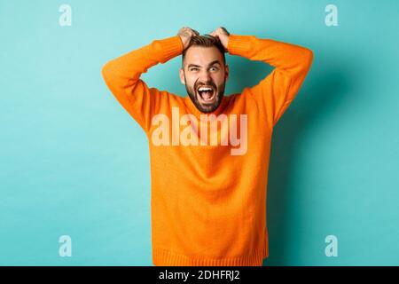Frustrated man shouting, pulling out hair and screaming angry, losing temper and looking mad, standing over light blue backgroun Stock Photo