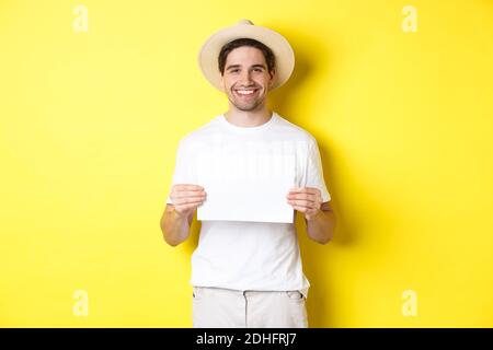 Handsome young male tourist in summer hat smiling, holding blank piece of paper for your sign, standing over yellow background Stock Photo