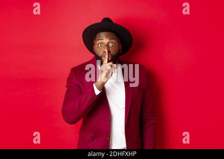 Fashion and party concept. African american man in classy hat hushing, asking keep secret, preparing surprise, standing over red Stock Photo