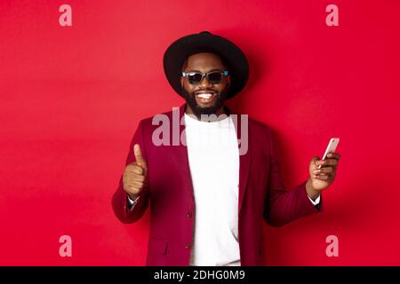 Handsome african american man using phone app and showing thumb up, smiling at camera, wearing sunglasses and fancy hat, red bac Stock Photo
