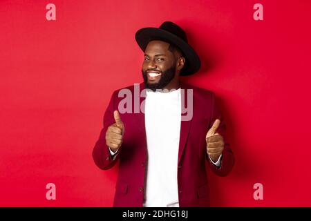 Cheerful Black man having fun on party, showing thumbs up in approval, smiling and liking something, standing against red backgr Stock Photo