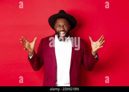 Angry Black man screaming and shaking hands with hatred, losing temper, standing outraged against red background Stock Photo