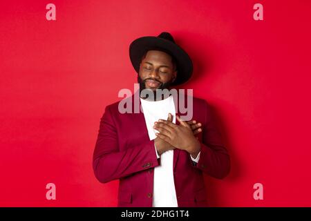Fashion and party concept. Young bearded Black man in classy hat and jacket, holding hands on heart and smiling nostalgic, remem Stock Photo