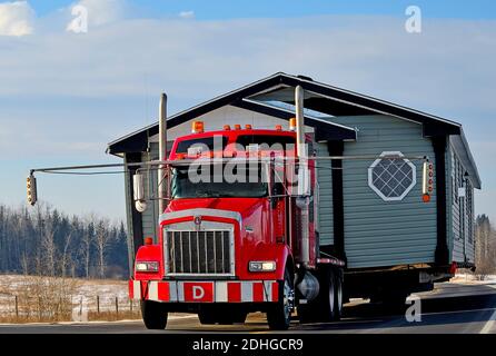 A close up image of a red semi truck moving a house along a two lane highway in rural Alberta Canada Stock Photo